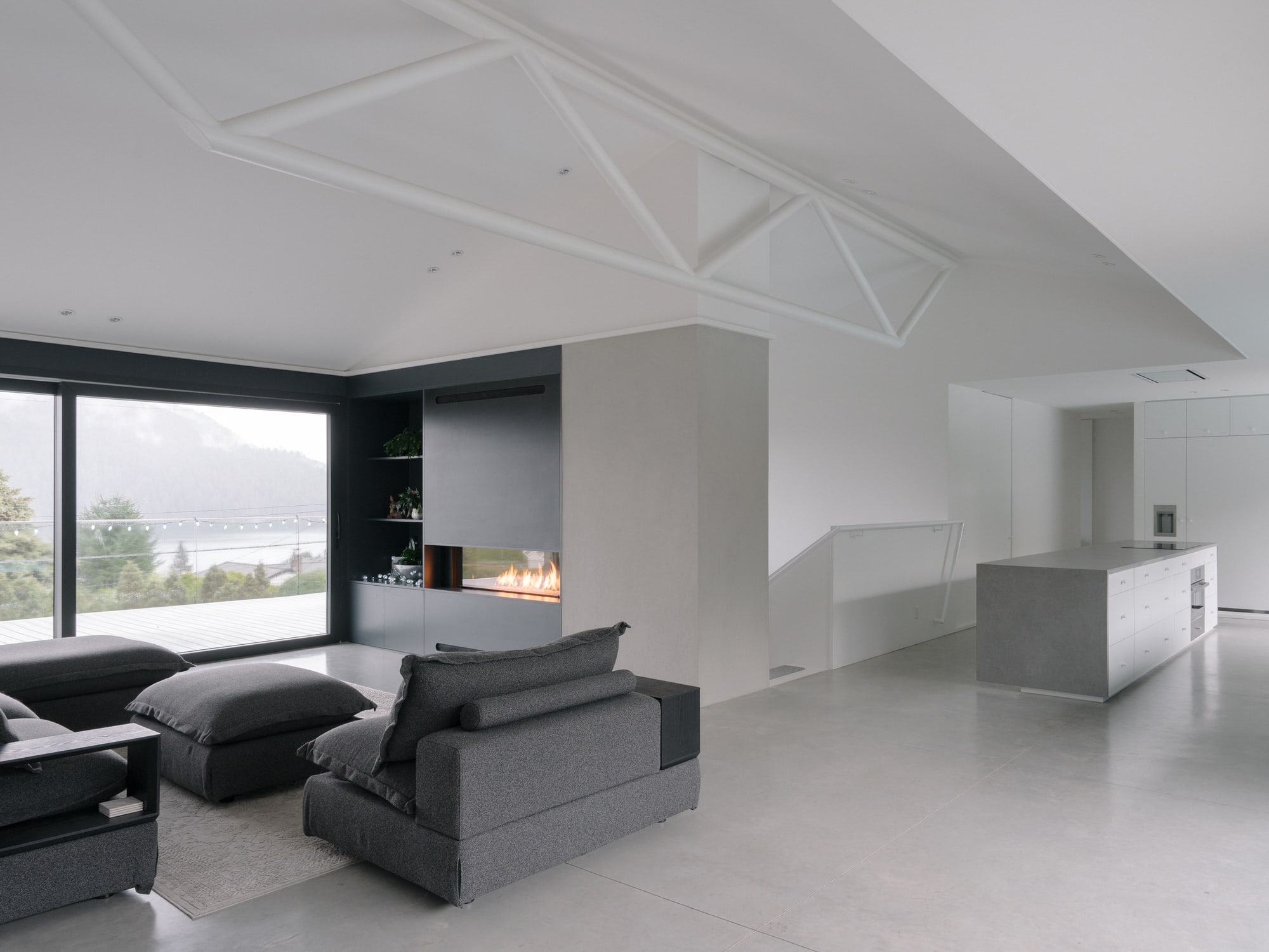 Baycrest — Open living room, fireplace and kitchen beneath white roof trusses